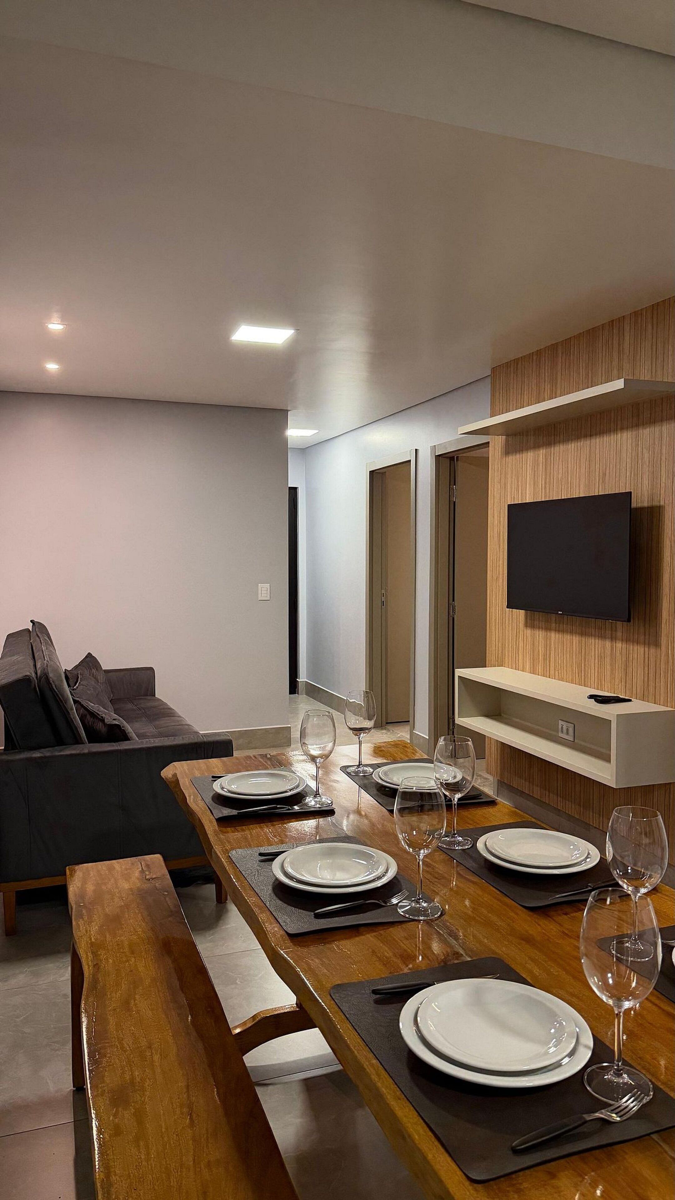 Uma área de refeições moderna com uma mesa de madeira comprida posta para seis pessoas, um sofá e uma TV fixada na parede.