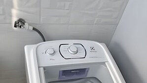 Een witte wasmachine in een hoek, met een snoer aangesloten, een blauwe emmer en een gele tas in de buurt.