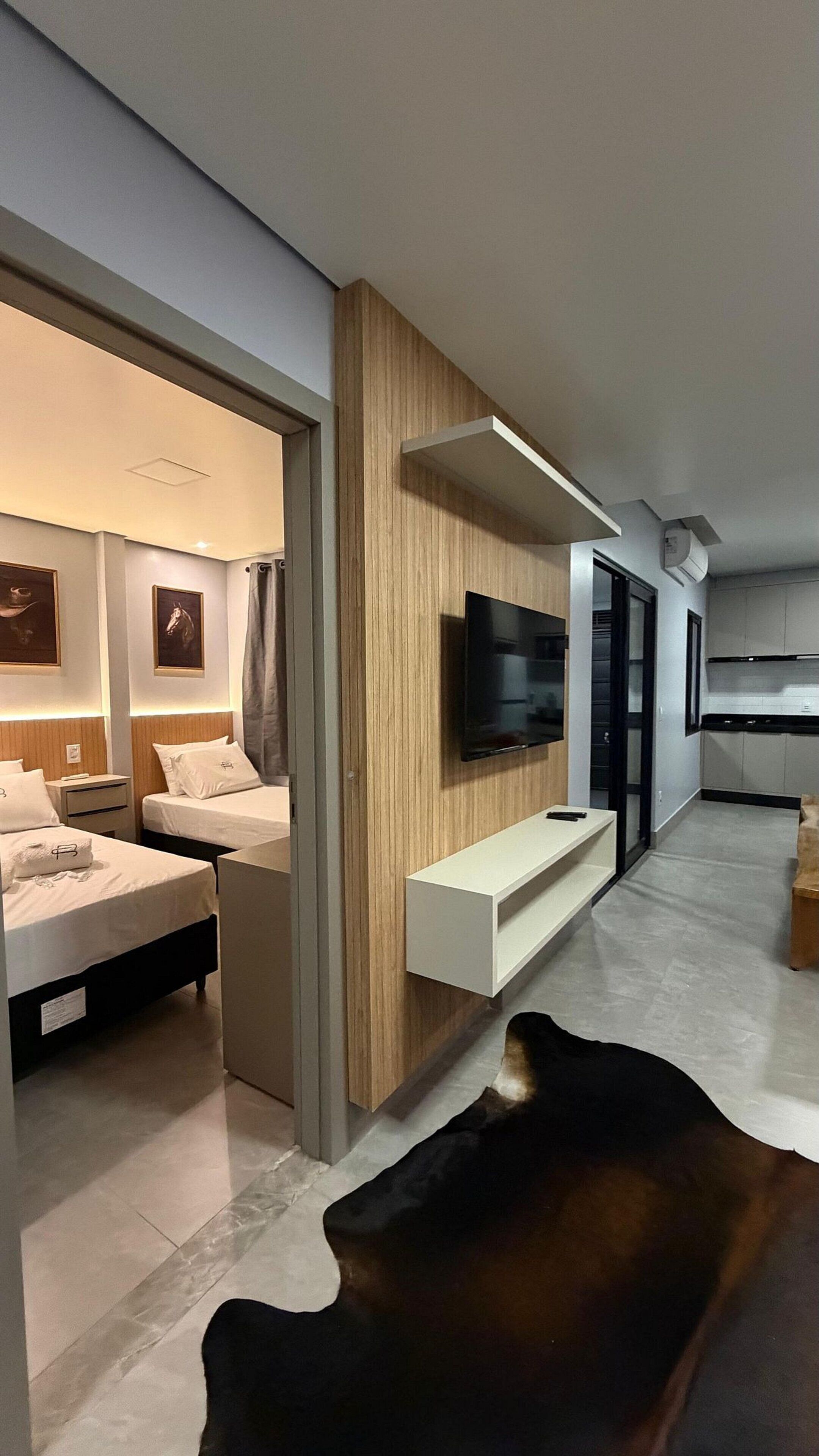 A modern hotel room with a flat-screen TV mounted on a wooden panel, a sofa, and a dining area with a table and chairs.