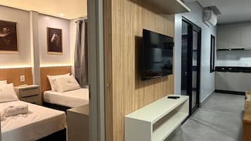 A modern hotel room with a flat-screen TV mounted on a wooden panel, a sofa, and a dining area with a table and chairs.