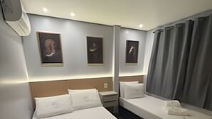 Hotellihuone, jossa on suuri sänky, pienempi sänky, yöpöytä, seinälle asennettu ilmastointilaite ja ikkuna, jossa on verhot.