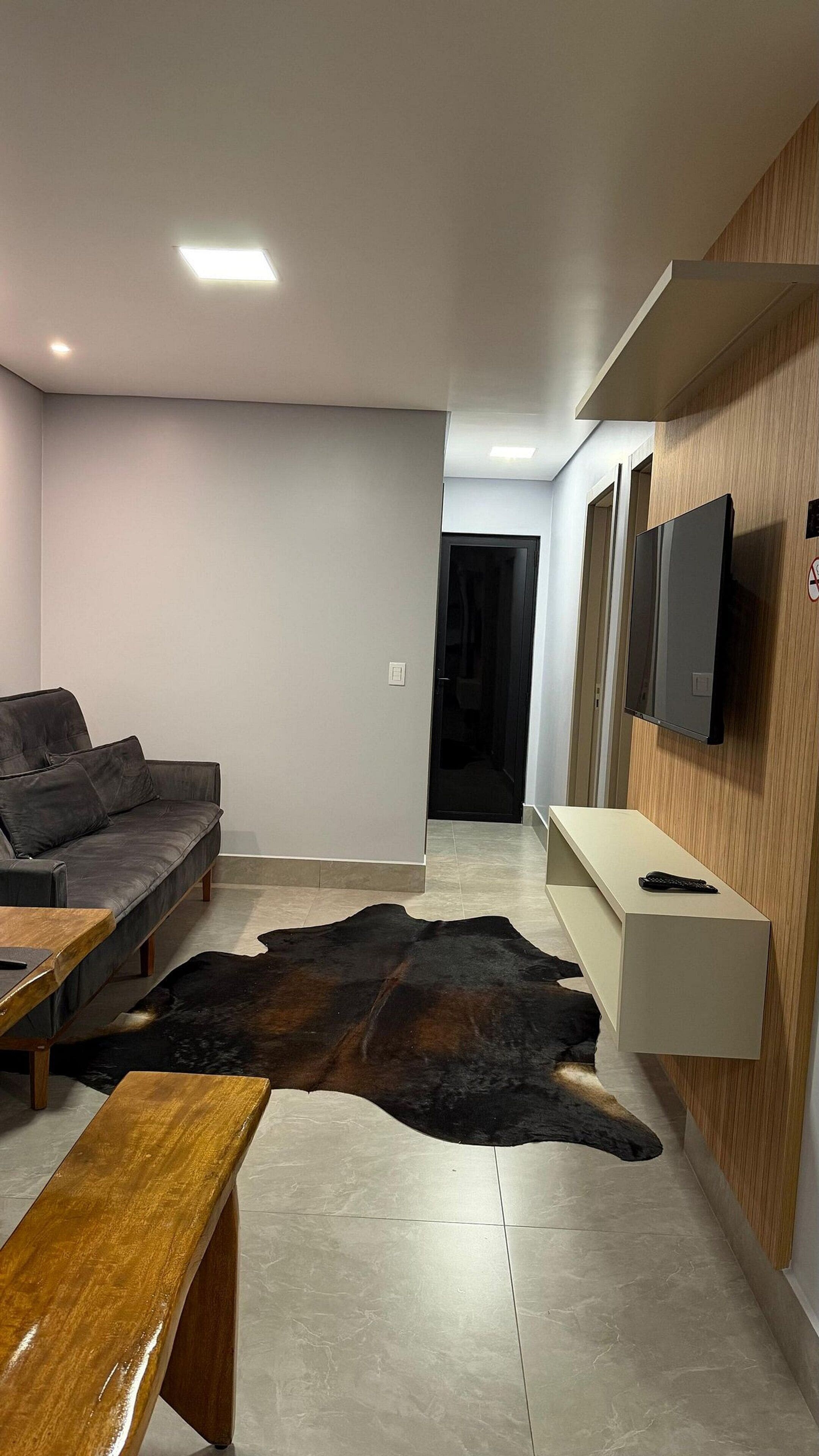 Uma sala de estar moderna com sofá, mesa de centro de madeira, tapete de couro e TV na parede.