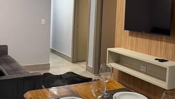 Un comedor moderno con una mesa de madera dispuesta para dos personas, un televisor de pantalla plana en la pared y un sofá gris al fondo.