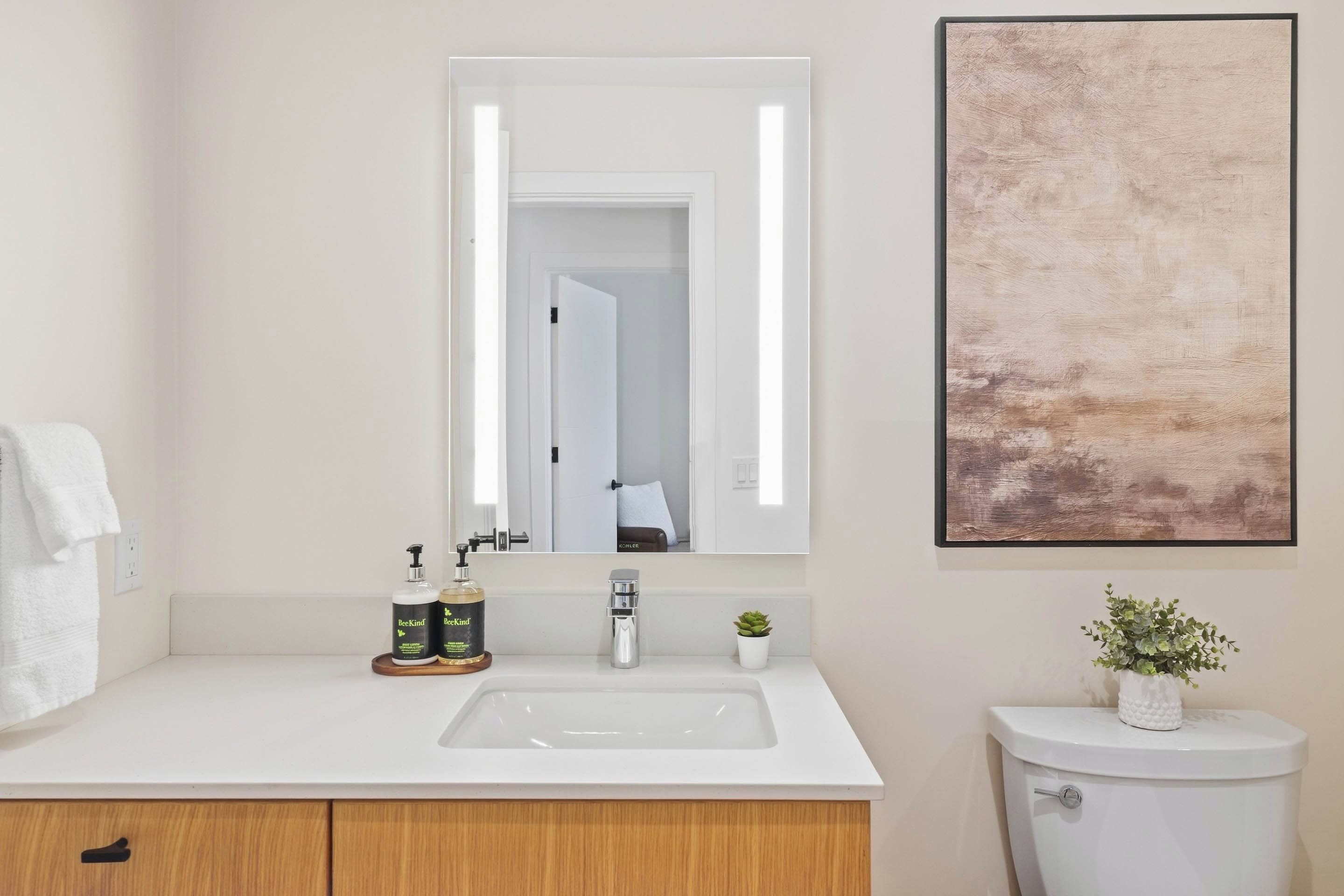 Une salle de bain avec un lavabo blanc, un miroir, une œuvre d’art encadrée, des toilettes et une petite plante en pot.