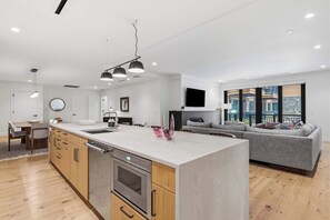 Eine moderne Küche mit einer zentralen Kochinsel, einem Essbereich mit Tisch und Stühlen, einem grauen Sofa und einem großen Fenster mit Blick auf die Stadt.
