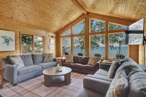 L’intérieur d’un chalet accueillant, avec un plafond en bois, de grandes fenêtres, un canapé gris, un divan en cuir marron et une petite table ronde.