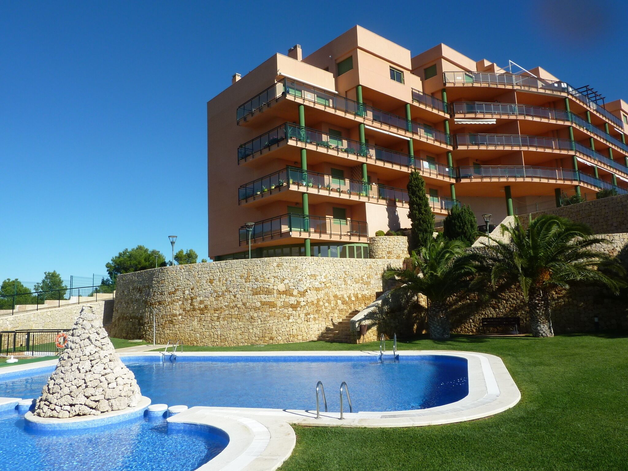A multi-story hotel with balconies, a swimming pool with a stone feature, and a stone wall.