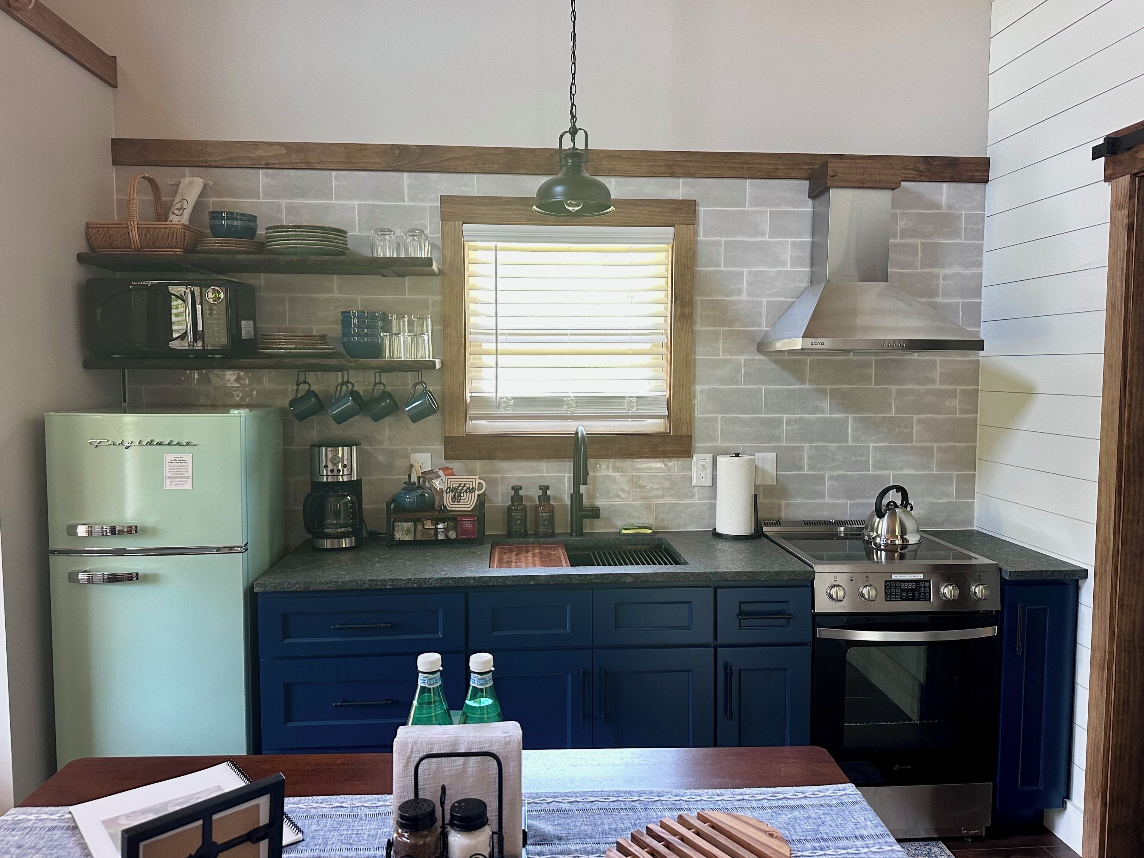 A kitchen with a vintage refrigerator, a coffee maker, a kettle, and a window with blinds.