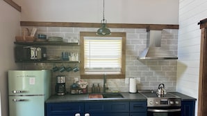 Une cuisine équipée d’un réfrigérateur vintage, d’une machine à café, d’une bouilloire et d’une fenêtre avec des stores.