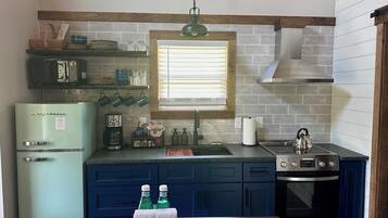 A kitchen with a vintage refrigerator, a coffee maker, a kettle, and a window with blinds.