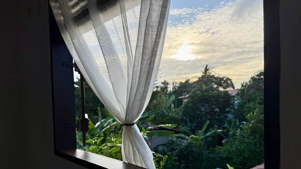 A window with sheer curtains, a view of greenery, and a building in the background. - Oceana Suites Veralucia (Tibau do Sul)