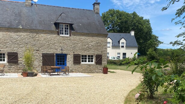 Una casa de piedra con camino de grava, una puerta azul y un pequeño patio con mobiliario exterior.