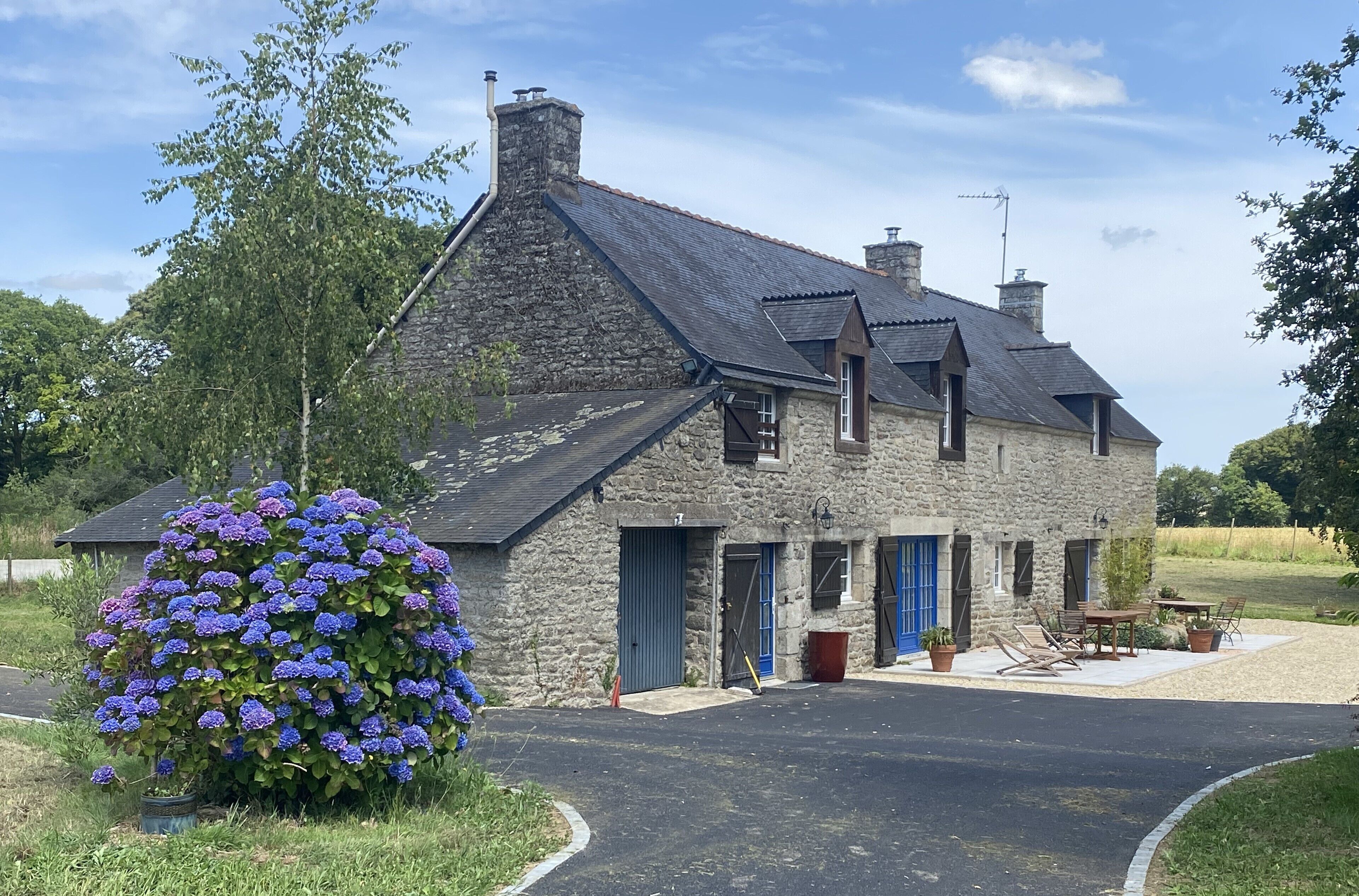 Une maison en pierre avec un toit en pente, un garage et une petite terrasse aménagée avec du mobilier d’extérieur.