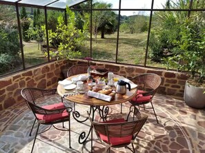 Une terrasse ensoleillée avec un sol en pierre, une table ronde dressée pour le petit-déjeuner, des chaises en osier avec des coussins rouges, et une vue sur un jardin luxuriant à travers de grandes fenêtres.