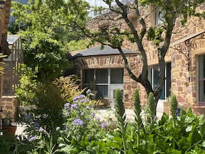 A stone building with a covered patio, surrounded by lush greenery and flowering plants.