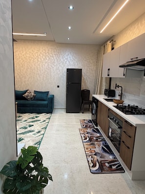 A modern kitchen with a refrigerator, stove, and sink. There is a dining area with a sofa and a decorative rug. - Steps to the Beach. NEW 1BR with Garage & Netflix (Kenitra)