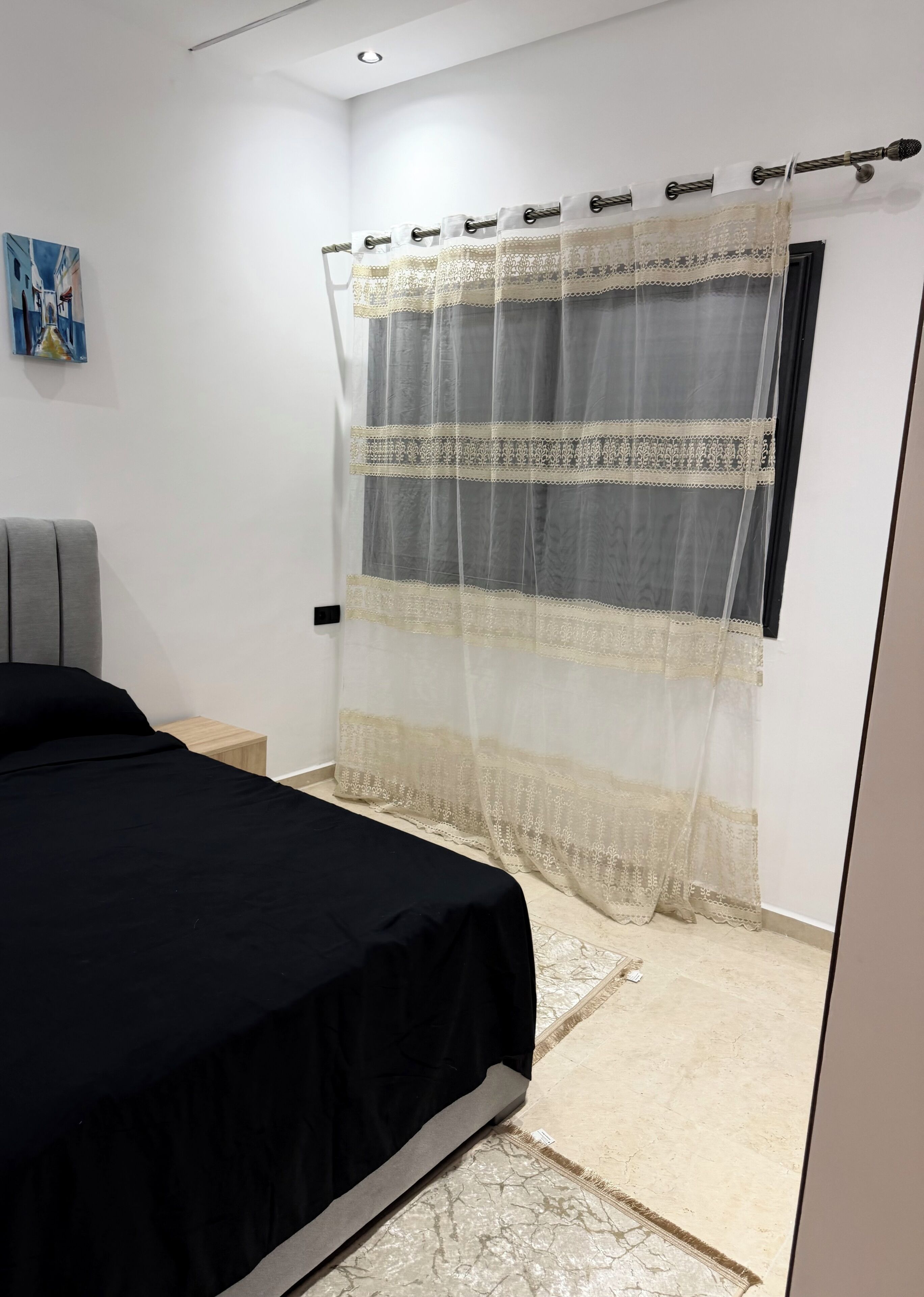 Een slaapkamer met een bed, een nachtkastje, een raam met gordijnen en een schilderij aan de muur.