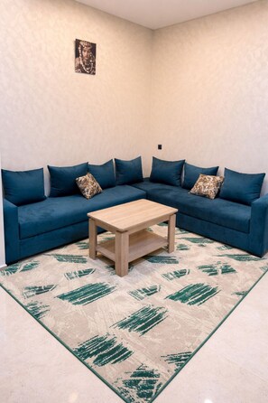 A corner living room with a blue sofa, a wooden coffee table, and a patterned rug. - Steps to the Beach. NEW 1BR with Garage & Netflix (Kenitra)