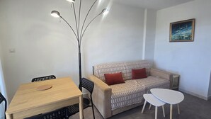 Ein Zimmer mit einem Holztisch, schwarzen Stühlen, einem beigen Sofa mit roten Kissen und einem weißen Couchtisch. An der Wand hängt ein Gemälde.