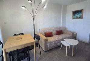A room with a wooden table, black chairs, a beige sofa with red cushions, a white coffee table, and a painting on the wall. - Studio for 4 with pool, 350m from beach (Fréjus)