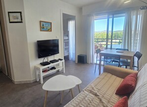 A living room with a flat-screen TV, a white coffee table, a sofa, and a dining area with chairs and a table. - Studio for 4 with pool, 350m from beach (Fréjus)