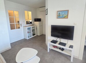 A compact living space with a kitchenette, a flat-screen TV mounted on the wall, a small dining table, and a white cabinet with glass doors. - Studio for 4 with pool, 350m from beach (Fréjus)