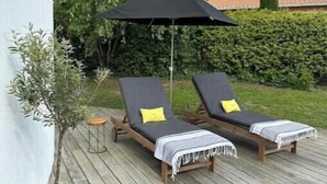 Ett trädäck med två solstolar och ett svart parasoll, omgivet av grönska och en vit vägg.