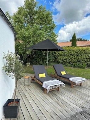 Um deck de madeira com duas cadeiras de descanso e um guarda-sol preto, cercado por vegetação e uma parede branca.