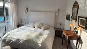 Ett sovrum med en stor säng, ett nattduksbord med en lampa, ett litet bord med en lampa och ett fönster med gardiner.