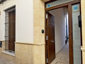 Ein Gebäudeeingang mit einer Holztür, einem vergitterten Fenster und einem mit Fliesen ausgelegten Weg.