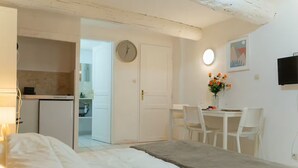 Ein Schlafzimmer mit einem Bett, einem Nachttisch, einem Schreibtisch mit Stühlen, einer Uhr, einem Fernseher und einer kleinen Küchenzeile.