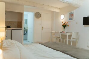 Ein Schlafzimmer mit einem Bett, einem Nachttisch, einem Schreibtisch mit Stühlen, einer Uhr, einem Fernseher und einer kleinen Küchenzeile.