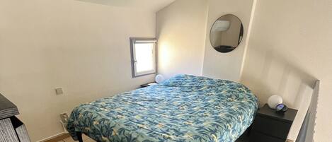 Un dormitorio con cama, una mesita de noche con lámpara, una ventana y un espejo en la pared.