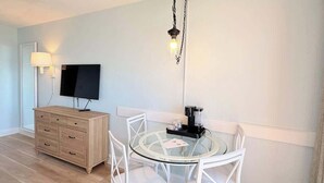 Ein Zimmer mit einem runden Glastisch, weißen Stühlen, einer Holzkommode, einem Flachbildfernseher und einer Pendelleuchte.