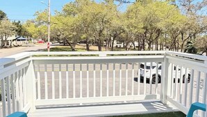 A porch with two blue chairs, a white railing, and a view of trees and a street. - San-Joy Retreat #9, 2BR Condo, Steps to Beach (Myrtle Beach)
