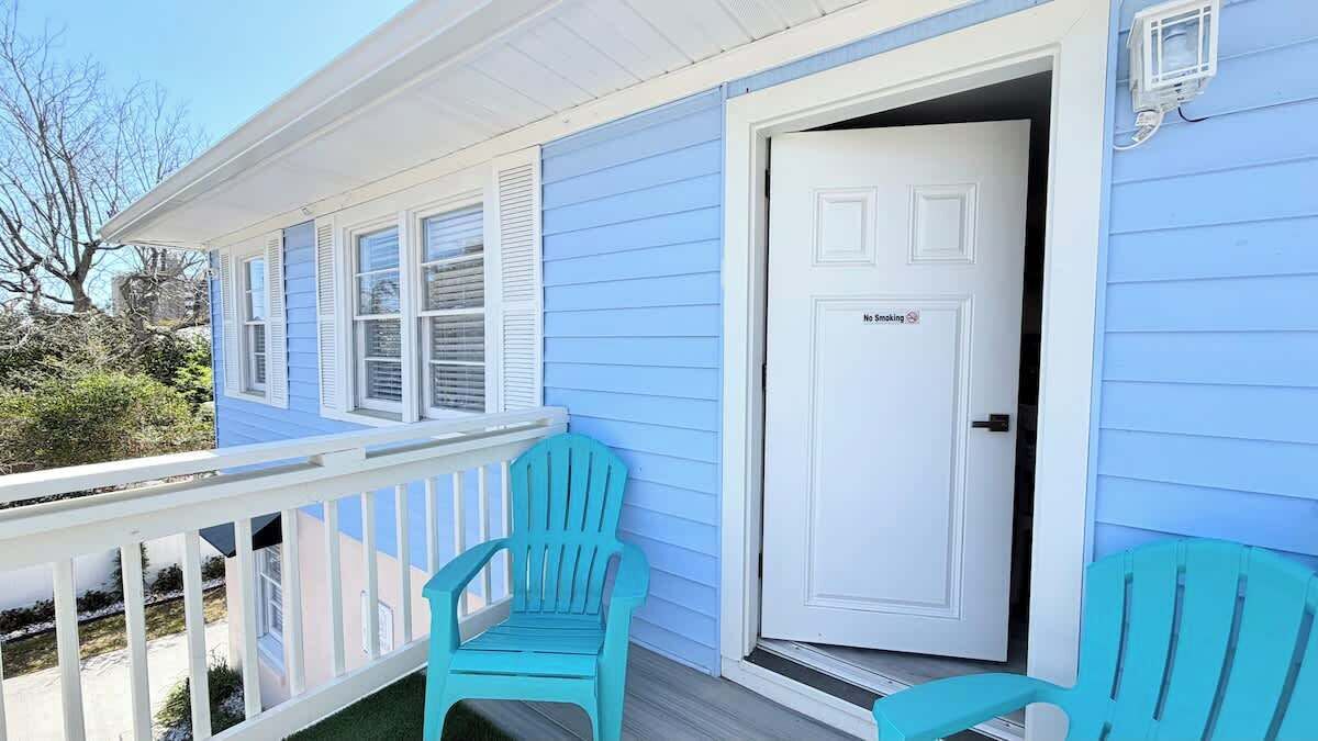 Une maison avec une porte blanche portant l’inscription « Il est interdit de fumer », une véranda avec des chaises turquoise et une balustrade blanche.
