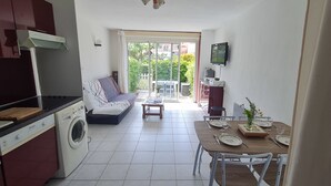 Cocina compacta con lavadora, comedor con mesa para dos, sofá y televisor montado en la pared.