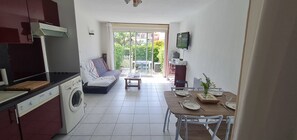 A compact kitchen with a washing machine, a dining area with a table set for two, a sofa, and a TV mounted on the wall. - Apartment with terrace & pool (Palavas-les-Flots)