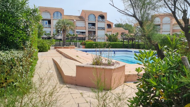 Un área de piscina con solárium de baldosas, rodeada de vegetación y un edificio con balcones.