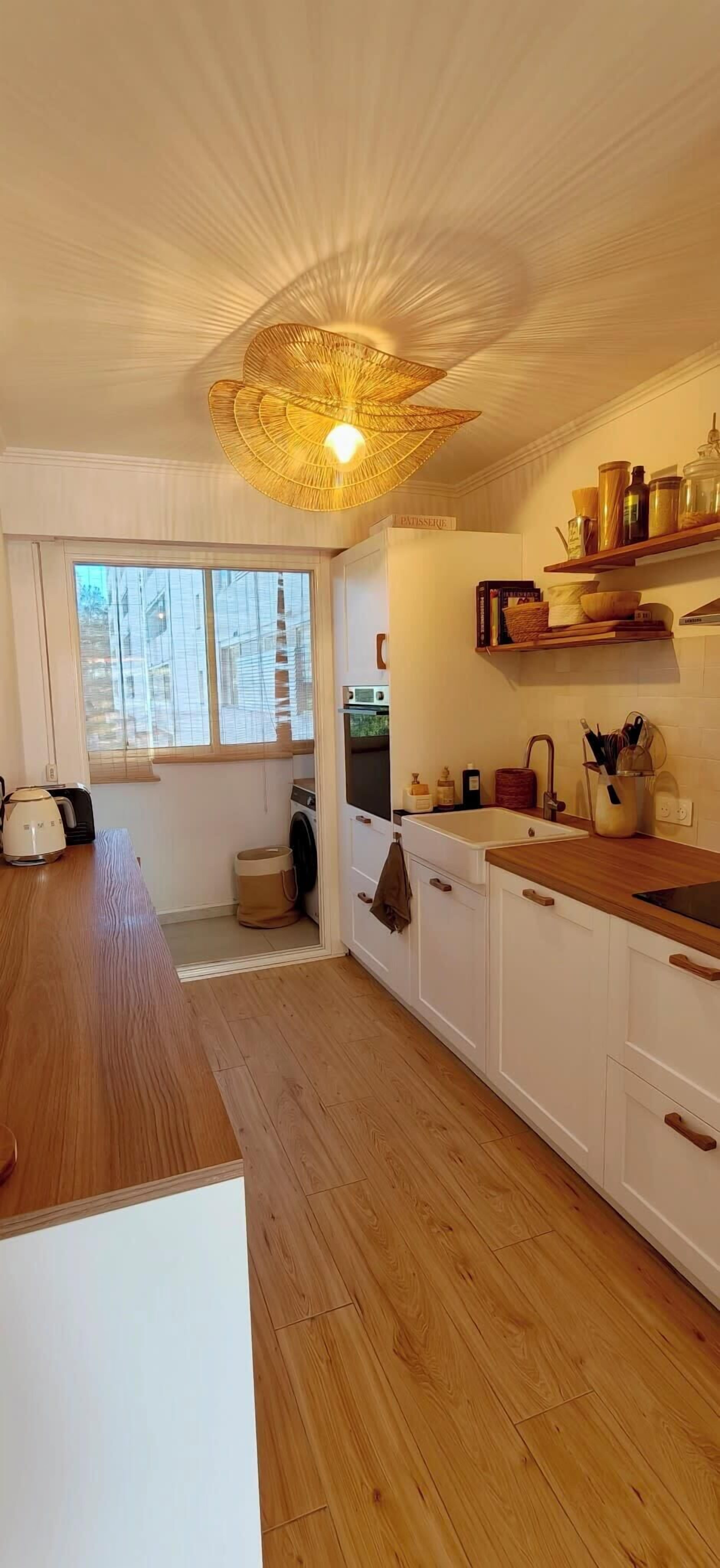 A modern kitchen with wooden flooring, white cabinets, a large window, and a unique ceiling light fixture.