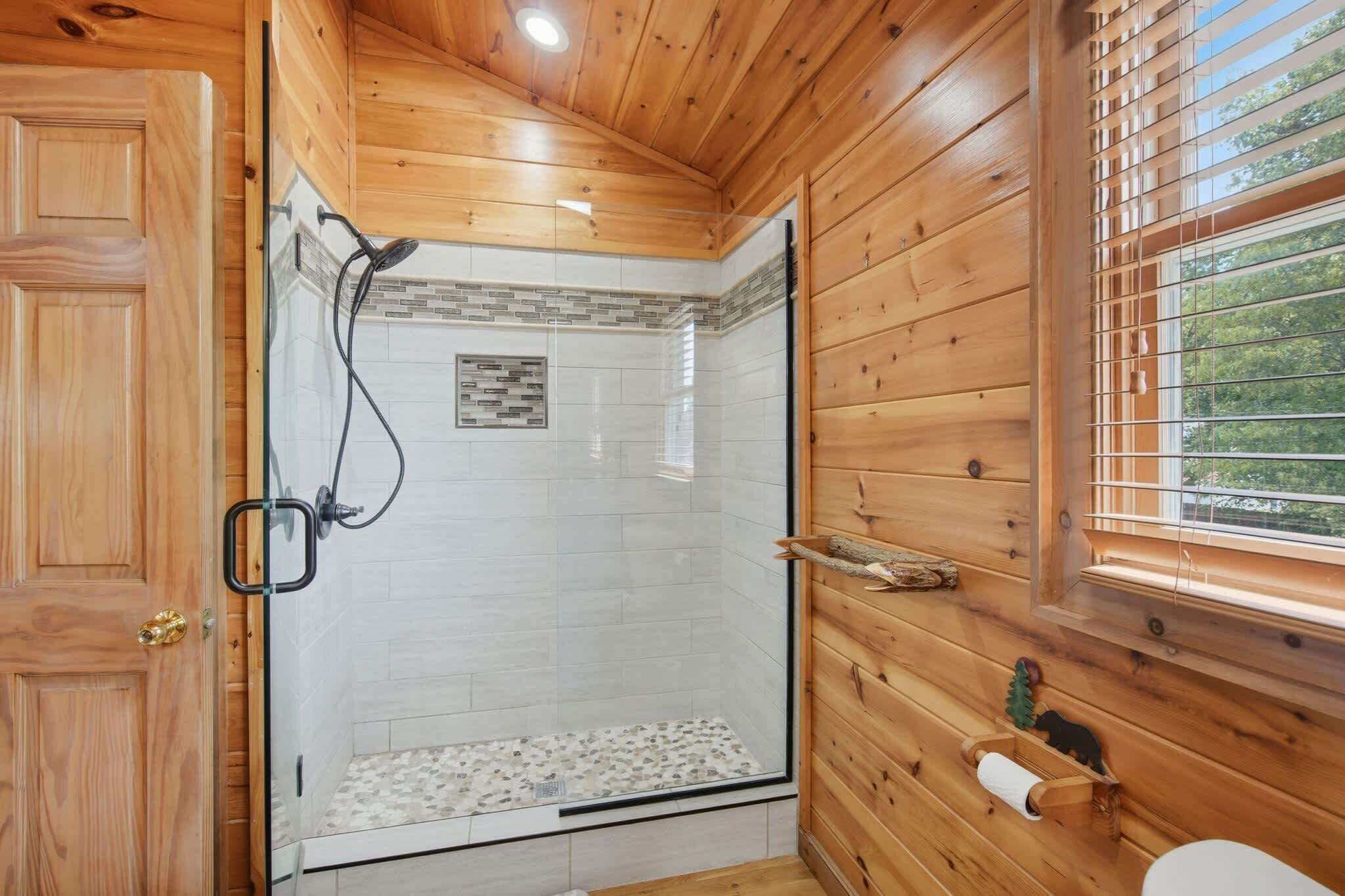 Une salle de bain rustique, avec des murs en bois, une douche en verre, des toilettes et une fenêtre équipée de stores.