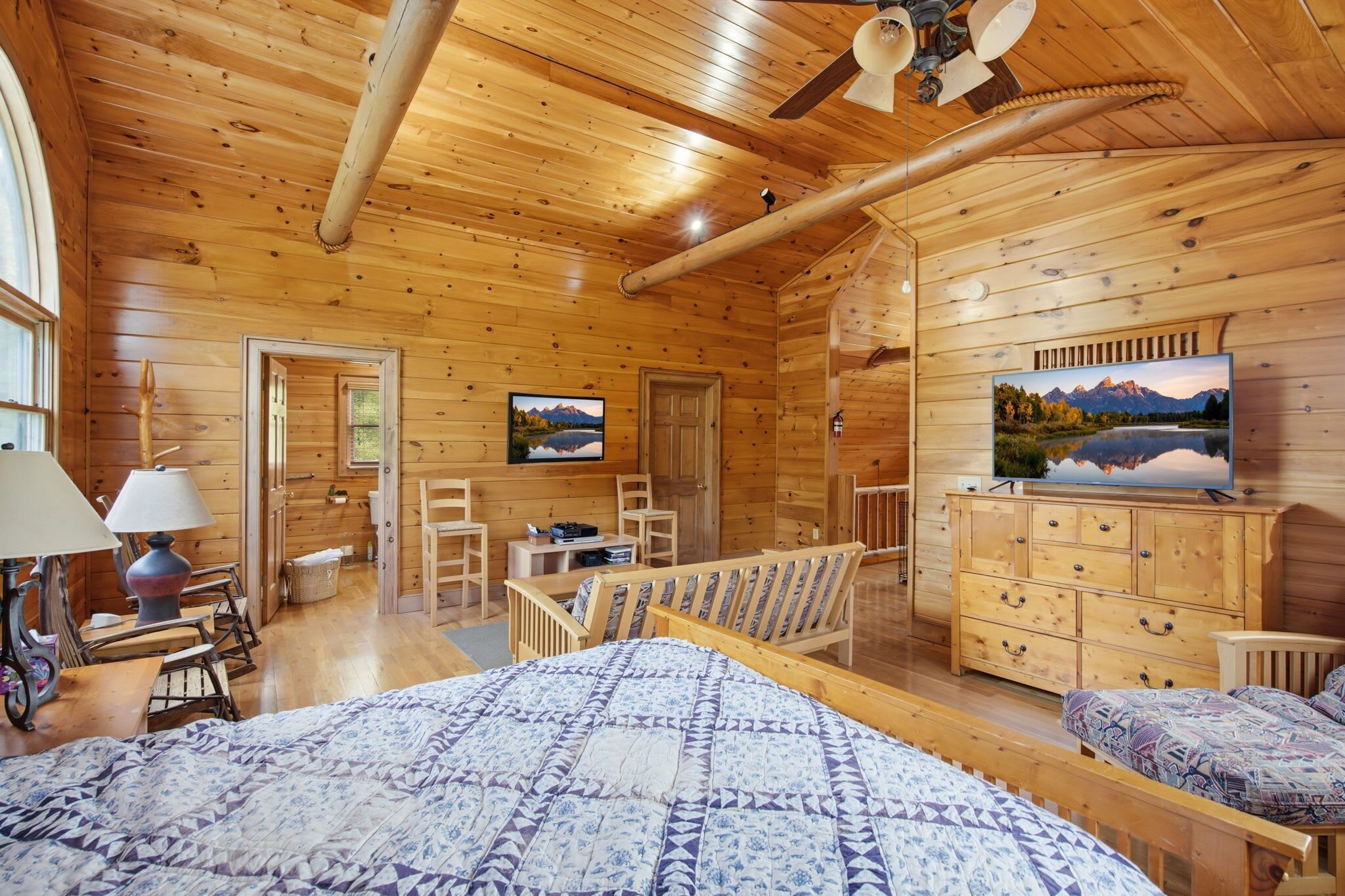 Interior de una cabaña de madera con una cama, televisor, escritorio y una silla.