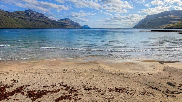 Una playa con aguas cristalinas, montañas a lo lejos y un cielo parcialmente nublado.