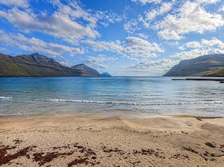 A beach with clear water, distant mountains, and a partly cloudy sky.. Beachhouse Syðrugøta