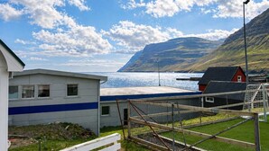 Un edificio frente al mar con una franja azul, un área cercada y una montaña al fondo.