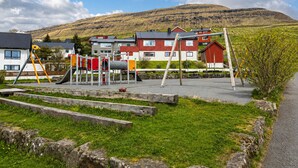 Un parque infantil con columpios y toboganes, un edificio rojo y blanco, y una colina al fondo.