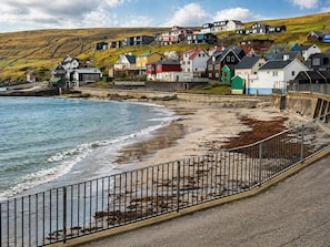 Un área residencial costera con casas a lo largo de la playa, una cerca que separa la playa de la carretera y un paisaje montañoso al fondo.
