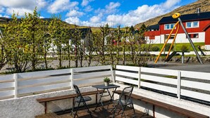Una terraza de madera con una mesa y sillas, barandas blancas y un área de juegos con un tobogán amarillo.