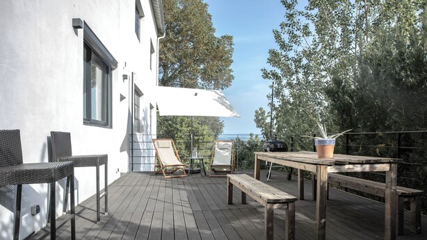 En träveranda med utemöbler, en vit byggnad och träd i bakgrunden.