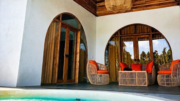 Un espace aménagé au bord de la piscine, avec des poutres en bois, des meubles en osier et une vue sur l’océan.